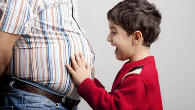 Father with striped shirt and visible belly, child in red sweater touches and smiles at the belly.