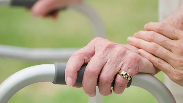 Elderly hands on walker, assisted by another person, symbolizing caregiving and support in a hospital setting.