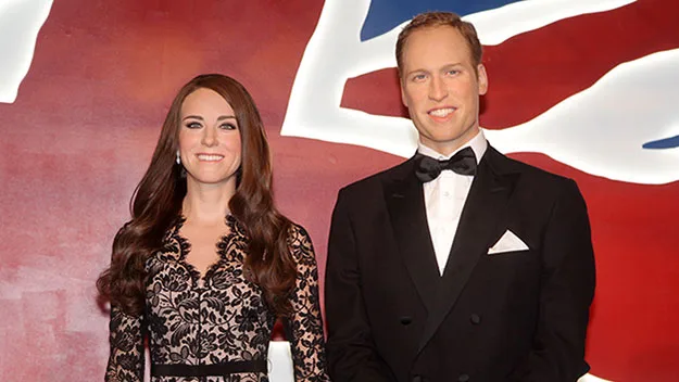 Wax figures of a woman in a lace dress and a man in a tuxedo, standing before a background resembling the Union Jack.