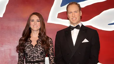 Wax figures of a woman in a lace dress and a man in a tuxedo, standing before a background resembling the Union Jack.