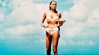 Woman in a white bikini holding shells on a beach, iconic scene from a classic 1962 movie under a blue sky.