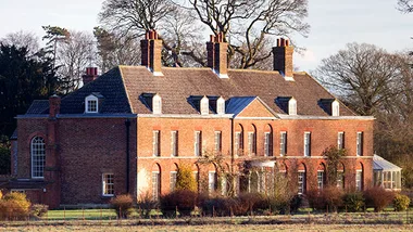 Red brick mansion with multiple chimneys, surrounded by trees and bushes, under a clear sky.