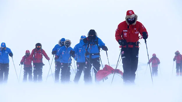 Skiers trek through a snowy landscape in Antarctica, wearing red and blue gear, with visibility low and wind blowing snow.
