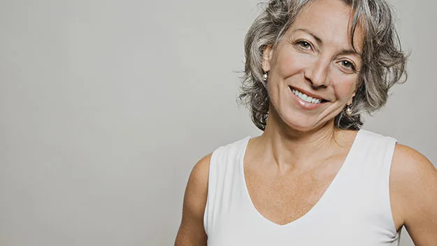 Smiling woman with gray hair in a white top against a neutral background.