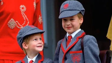 Two young boys in matching school uniforms with grey caps and red ties, smiling at each other.