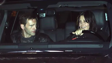 Two people sitting in a car at night, one driving and the other in the passenger seat, engaged in conversation.