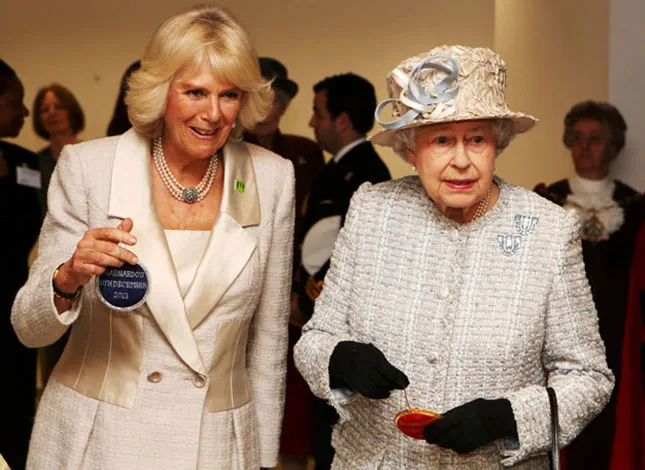 Royalty in formal attire: Woman in cream suit holds medal; another in a hat and gloves holds small purse, both smiling indoors.
