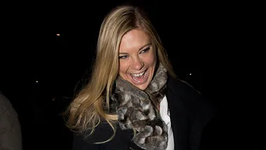 A smiling woman with long blonde hair, wearing a fur scarf and dark coat, captured at night.