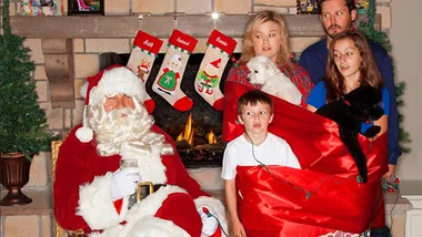 Family awkwardly poses with Santa, holding dogs in front of a fireplace with stockings, one child wrapped in red.