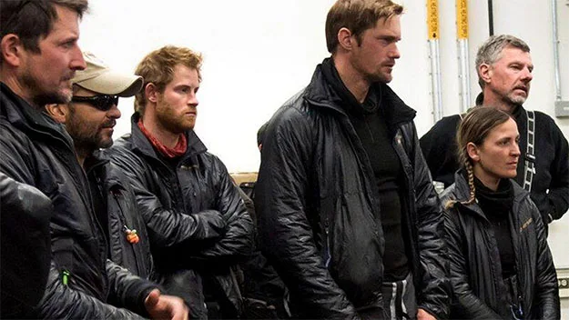 Men and women in outdoor gear, one with a red beard, stand indoors attentively, suggesting a group expedition.