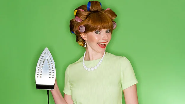 Woman with curlers and an iron, smiling against a green background, wearing a pearl necklace.