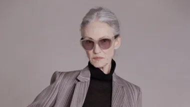 Elderly woman in striped suit and sunglasses modeling; dressed in dark turtleneck, posing with confident expression.