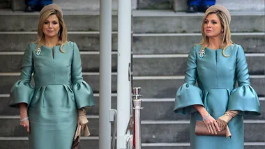 Queen Máxima in a teal dress with flared sleeves, wearing gloves and a matching hat, standing on stone steps.