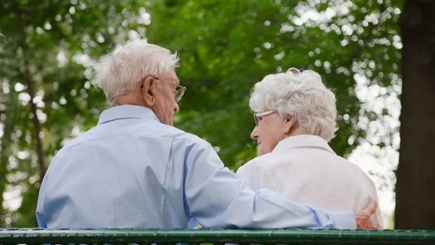 Elderly couple sitting closely on a bench outdoors, surrounded by greenery, sharing a tender moment.