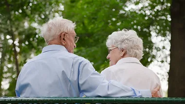 Elderly couple sitting closely on a bench outdoors, surrounded by greenery, sharing a tender moment.