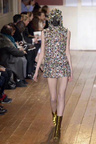 Model in a colorful gem-studded dress and mask walking down a wooden runway, with audience seated on both sides.