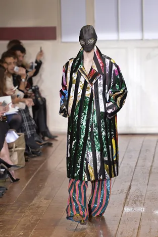 Model on runway wearing a colorful, patterned coat and pants with a black mask and goggles, audience observing.