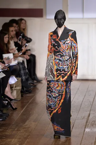 Model in vibrant, abstract-patterned suit and mask walks runway; audience watches in background.