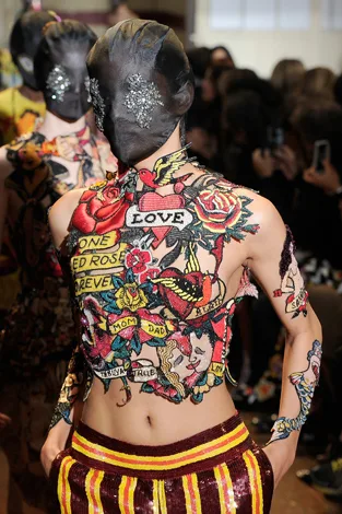 Fashion model in tattoo-patterned outfit and mask with floral designs walks runway at a fashion show.