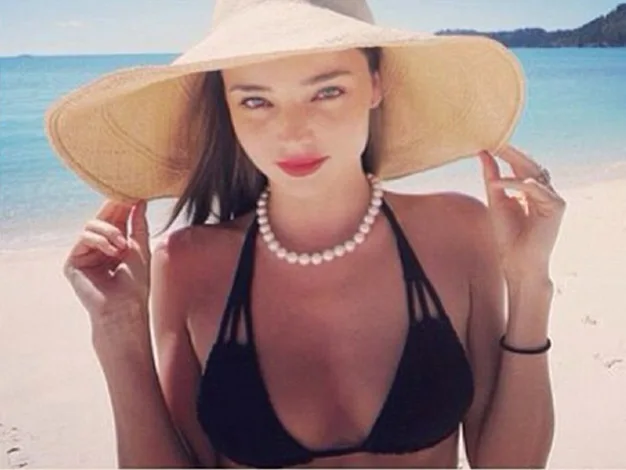 Woman in a black bikini, pearl necklace, and sun hat on a beach.