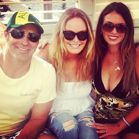 Three people sitting together, smiling, wearing sunglasses; one is in a cap, the others have long hair.