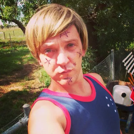 A person with short blond hair, wearing a tank top, has drawn marks on their face, standing outdoors near a fence.