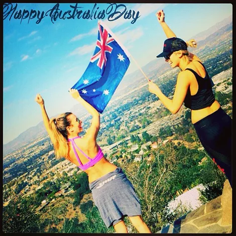 Two women celebrate outdoors holding an Australian flag, with a cityscape in the background. Text reads "Happy Australia Day."
