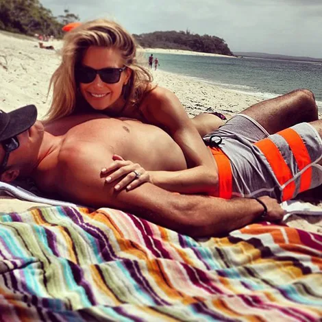 A couple on a beach, with a woman leaning over a man on a colorful striped towel. Both wear sunglasses, appearing relaxed.