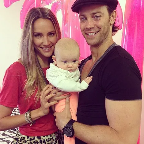 A couple smiling and holding a baby in a casual setting with pink wall art in the background.