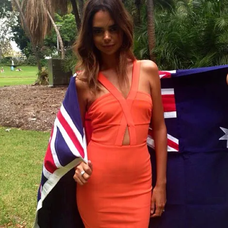 A woman in an orange dress stands outdoors holding an Australian flag, surrounded by greenery.