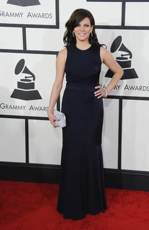 Woman in black gown holding silver clutch on red carpet at Grammy Awards.