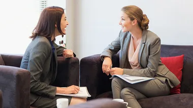 Two women in business suits having a meeting on a sofa, smiling and holding documents.