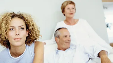 Three people in a relaxed setting; a young woman in focus, with an older couple in the background.