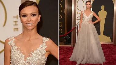 Woman in an embellished gown poses on the Oscars red carpet, smiling confidently with a hand on her hip.