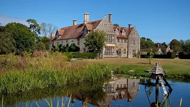 Medieval-style estate with red roof, surrounded by lush garden and pond, clear blue sky reflecting on water.