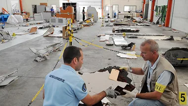 Debris from an aircraft laid out in a hangar with two investigators examining the pieces.