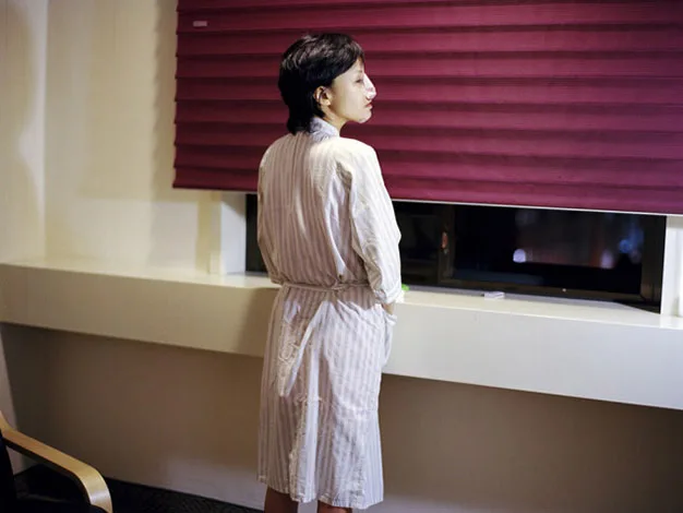 A person in a striped robe stands by a window with closed red blinds, looking outside.
