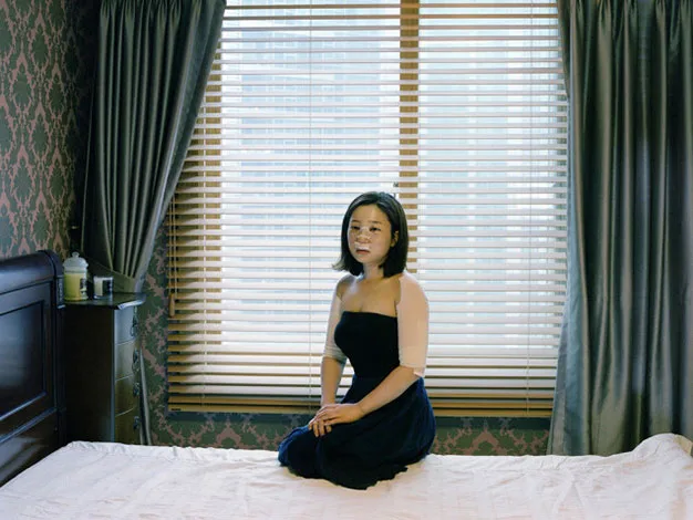 A woman in a dark dress sits on a bed in a dimly lit room with patterned wallpaper and large window blinds.