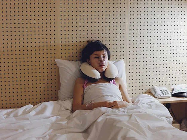 Person in bed with neck pillow and ice packs on cheeks, in a room with wood panel wall, phone on bedside table.