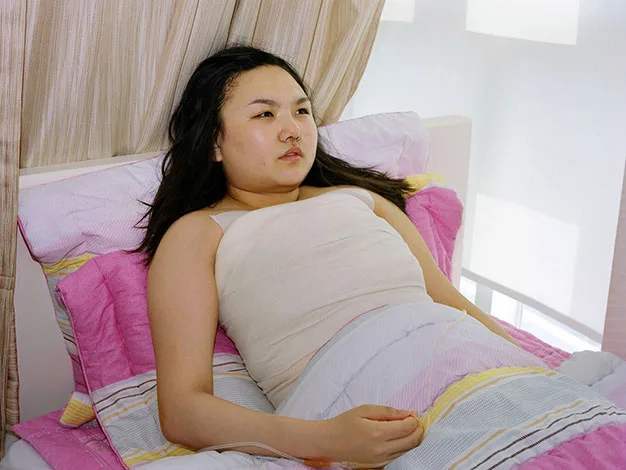 Young person lying in bed with a bandaged torso, gazing upwards, surrounded by pink and striped bedding.