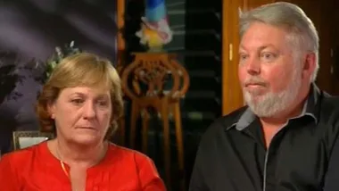 A woman in a red shirt and a man in a black shirt sit together indoors, looking serious.