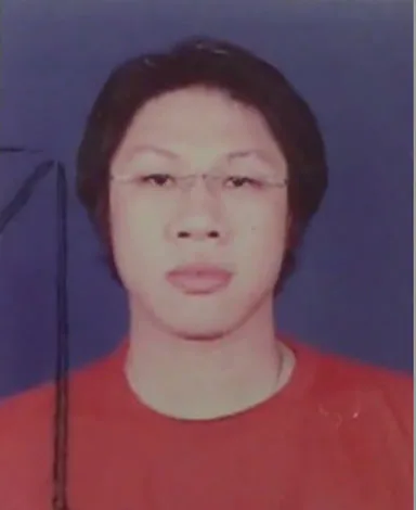 A passport-style photograph of a person with glasses and short dark hair, wearing a red top against a blue background.
