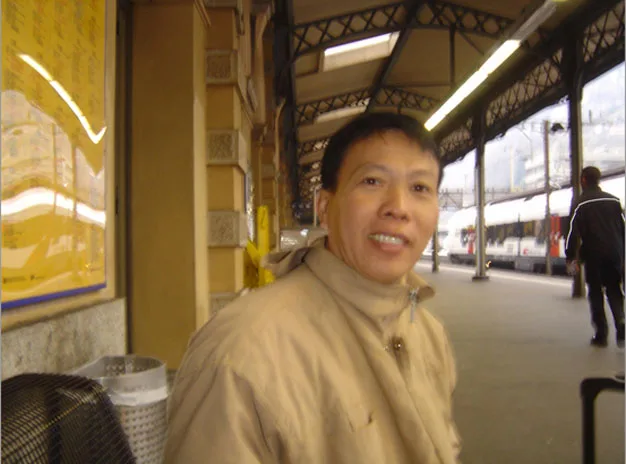 Person in a beige coat sitting at a train station platform with tracks and blurred figures in the background.