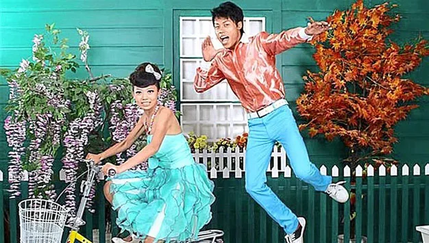 Man poses energetically near woman on bike in colorful vintage attire, set against vibrant plants and green backdrop.