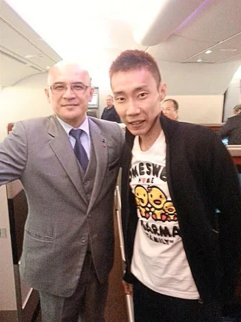Two men posing together in an airplane cabin, one in a suit and the other in a casual t-shirt and jacket.