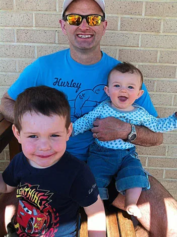 Father wearing sunglasses, seated with a smiling baby on his lap and a young child standing nearby, in casual attire.