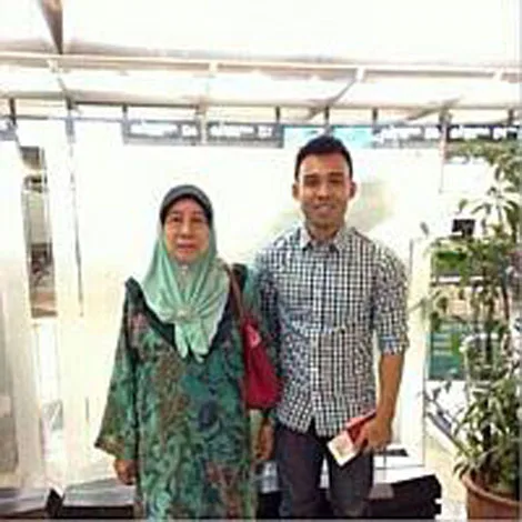 Two people stand side by side; the woman wears a hijab and floral dress, the man wears a checkered shirt, indoors.