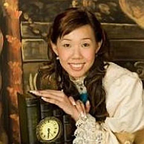 Young woman smiling, wearing a white blouse, posing with a vintage-style clock and bookshelf as a backdrop.