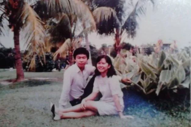 Couple sitting on grass in a garden with palm trees, smiling at the camera.