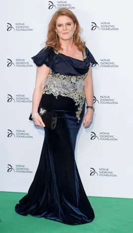 A woman in a blue gown with silver embellishments poses at an event, standing on a green carpet with a branded backdrop.